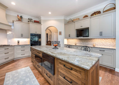 Kitchen renovation with view of island, stove, and sideboard, with leathered granite countertop, flush-set custom cabinetry, warming oven, microwave, finger mosaic tile backsplash, and multi-piece crown molding.