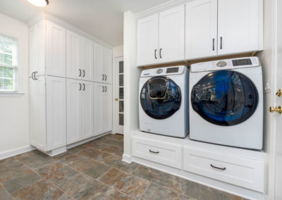 Renovated laundry room with view of raised, inset washer and dryer, white custom cabinetry with oil-rubbed bronze hardware, and tile floor.