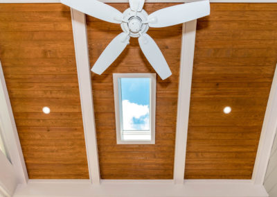 Porch renovation detail of tongue and groove vaulted ceiling, recessed lights, skylight, ceiling fan, faux beams, white trim, and contrasting color.