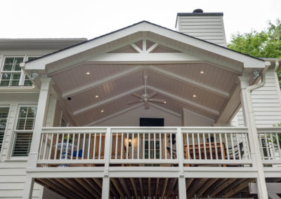Porch renovation with view from yard (centered) of railings, crowned corner posts, vaulted tongue and groove ceiling, recessed lighting, ceiling fan, exposed beams, and custom gable pediment design.